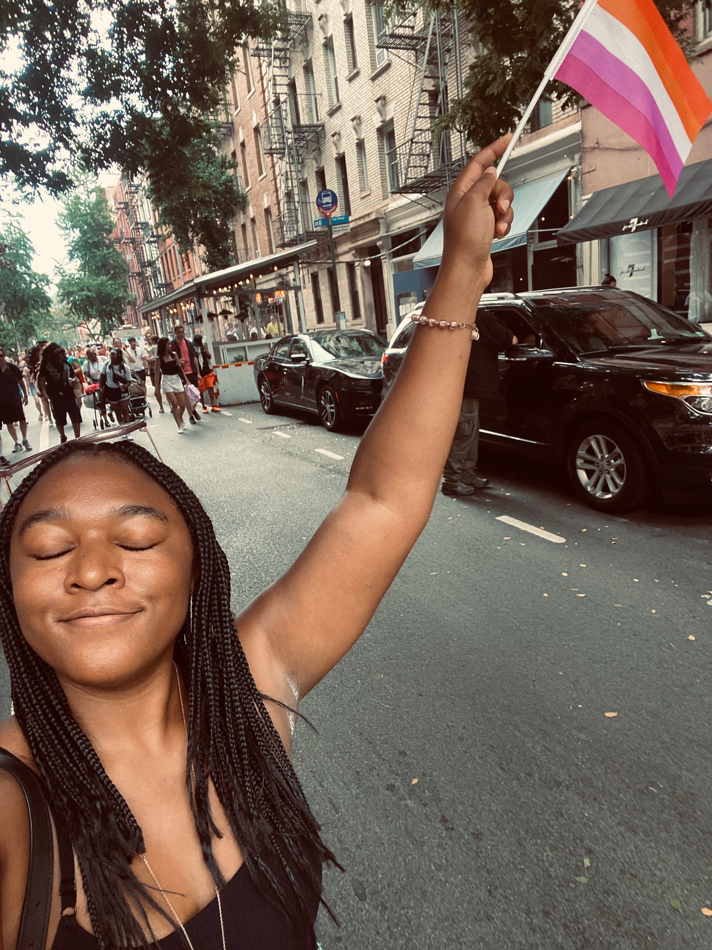 Alexa is holding up the lesbian pride flag (even though she identifies as bi) in the middle of a closed street in the west village. her eyes are closed and she is smiling peacefully. there is a crowd of people a few feet behind her.