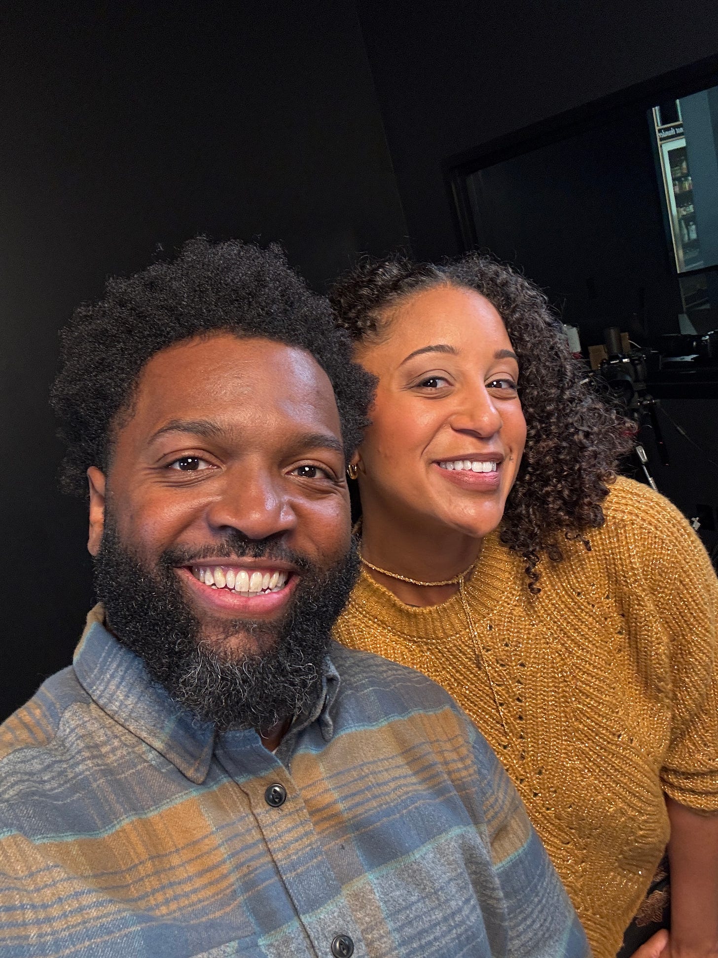 Two people smiling for a selfie in a warmly lit room. The man on the left has a beard and is wearing a plaid button-up shirt. The woman on the right has curly hair and is wearing a mustard yellow knit sweater. They appear happy and are standing close together.
