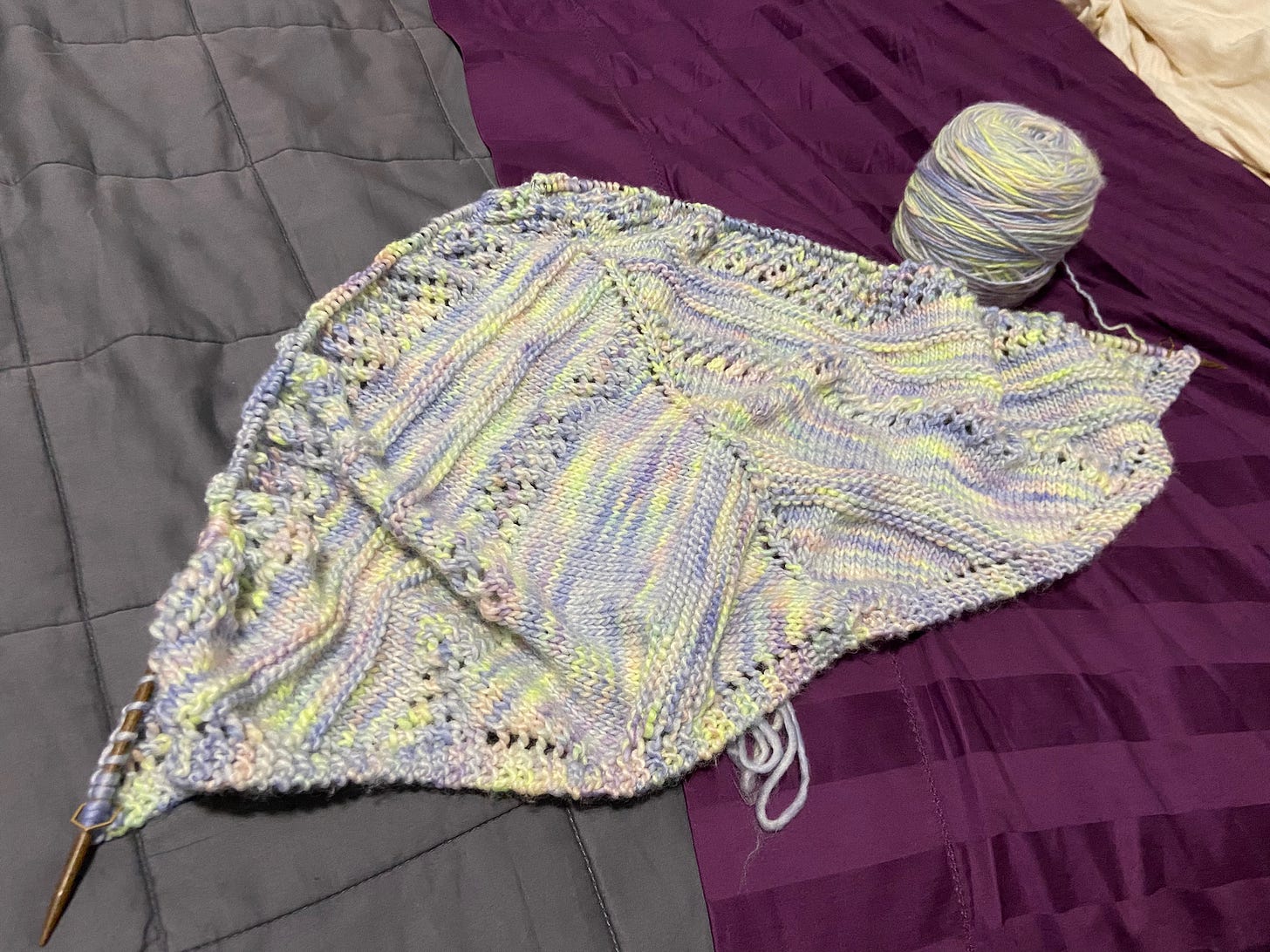 An in-progress Flying Fox Shawl laying on my bed. The yarn is in variegated blue, purple, green, yellow, and pink.