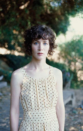 Photograph of MIranda July. A white woman with curly brown hair wearing a yellow dress with black spots. She is outside, standing in front of trees in daylight