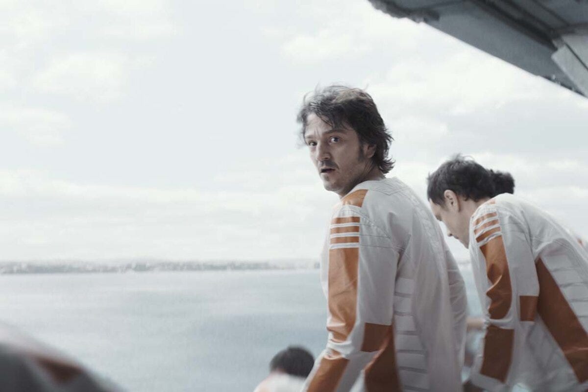 A man with unkempt brown hair, wearing a white and orange jumpsuit, looks at the camera as other men weraing the same clothes jump down a ledge