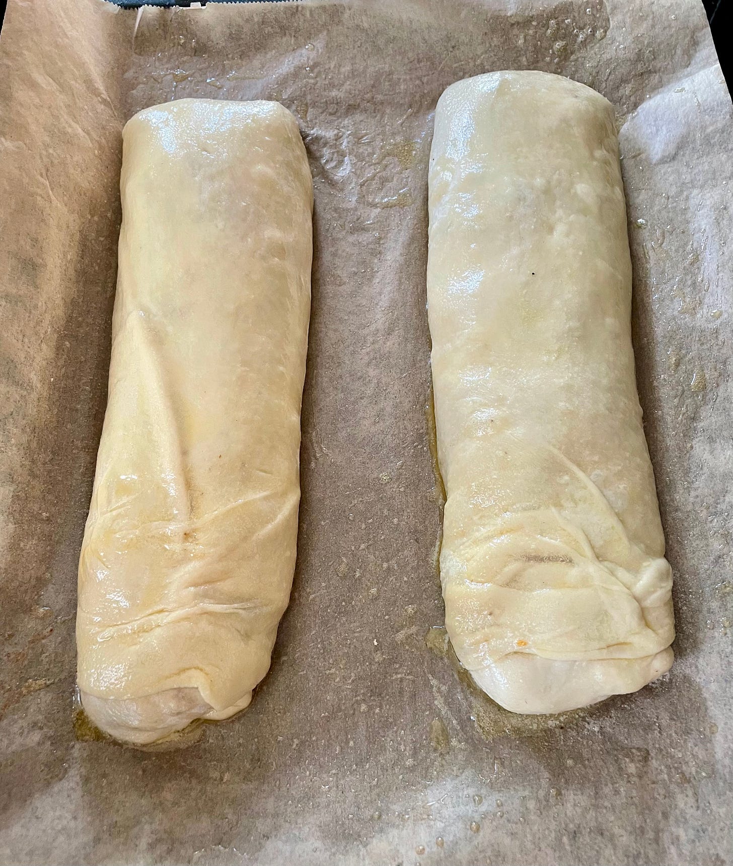 Two unbaked strudels placed on a parchment-lined baking tray, brushed with melted butter, ready for baking. Two unbaked strudels placed on a parchment-lined baking tray, brushed with melted butter, ready for baking.