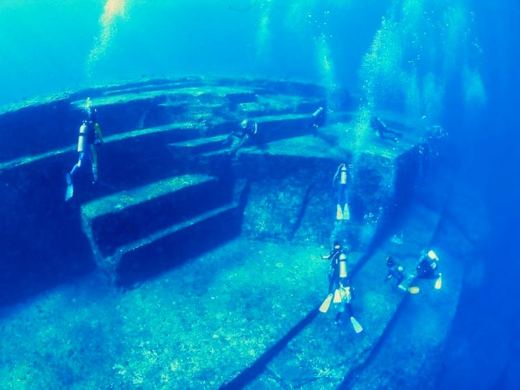 An underwater photo of divers exploring the Yonaguni Monument, a large rock formation with multiple very flat stones at various angles, including some that look like enormous stairs.