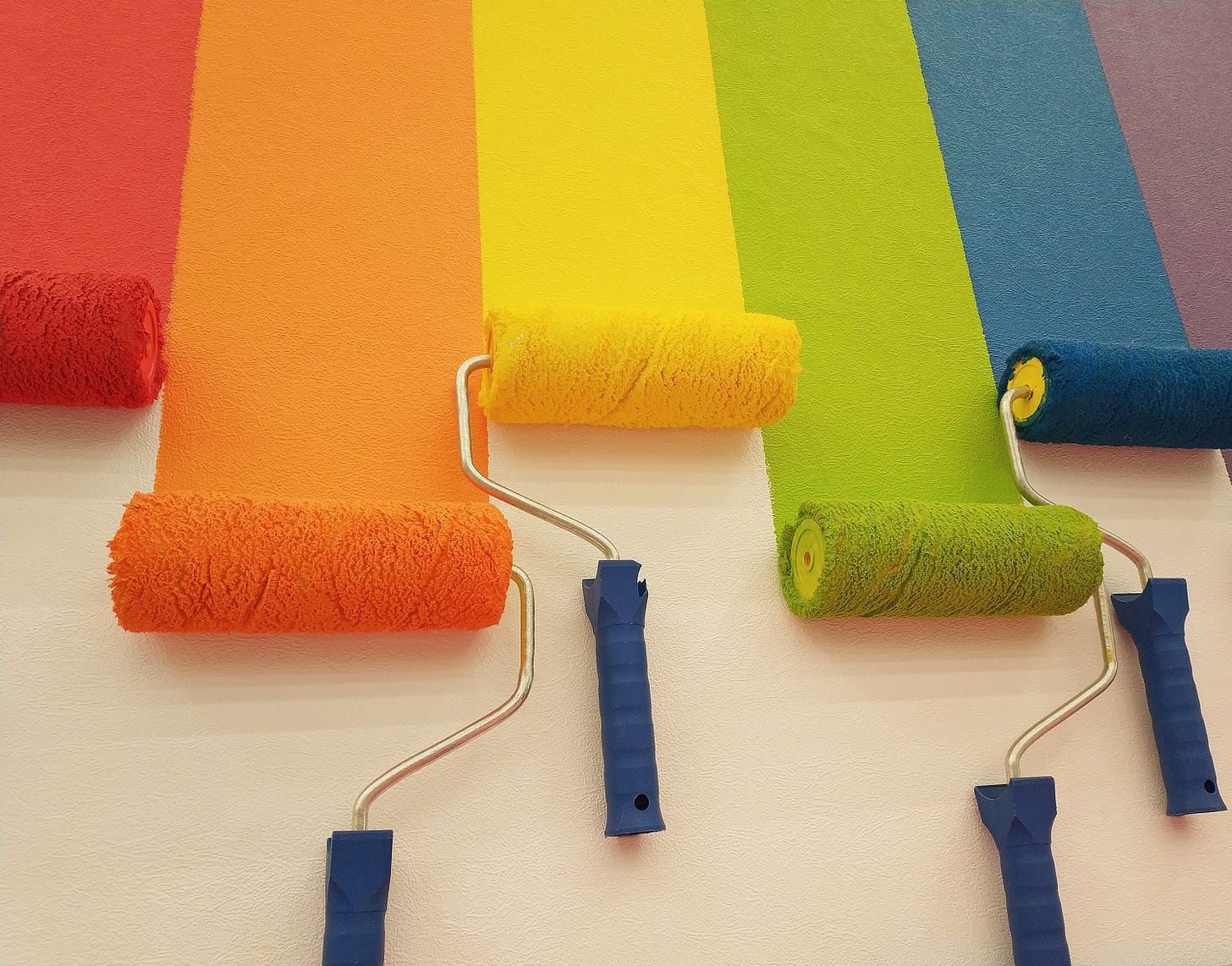 Six roller brushes side by side, painting a wall in colorful stripes: red, orange, yellow, green, blue, purple.