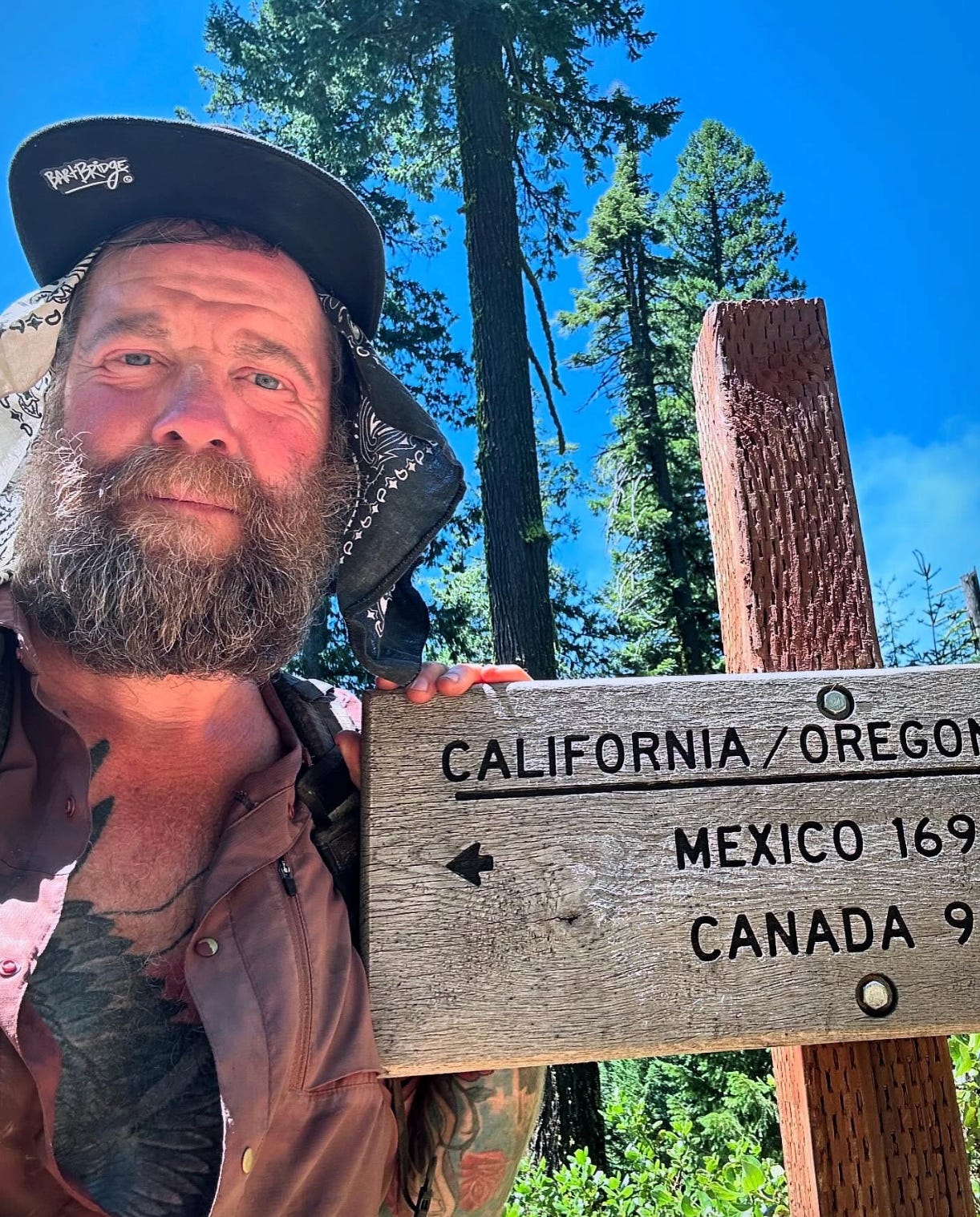 A man with a bushy beard next to a wooden sign marking the California/Oregon board; below, the signs points south toward Mexico and north toward Canada