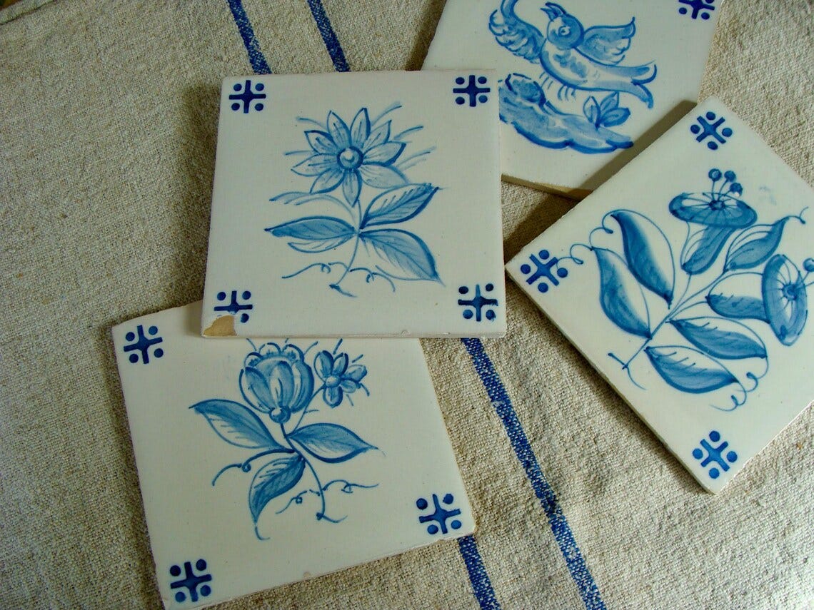 May include: Four square blue and white ceramic tiles with decorative floral and bird motifs.  The tiles feature hand-painted designs, including stylized flowers, leaves, and a bird.  The color palette is primarily blue and white, with the designs exhibiting a vintage or antique aesthetic. Each tile has a cross-shaped design in the corners. These decorative tiles are suitable for various craft projects or home decor applications.