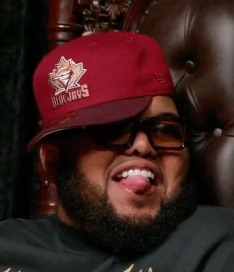 This may contain: a man wearing a maroon hat and glasses sitting in a chair with his tongue out