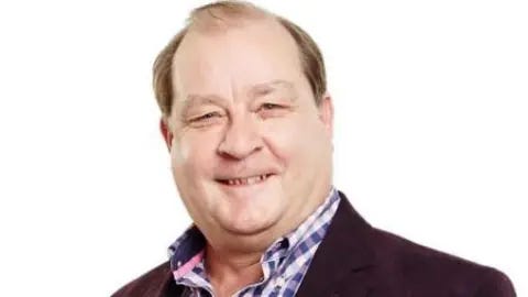 BBC Andy Ball with brown hair smiling wearinga pink and blue checked shirt and dark brown blazer