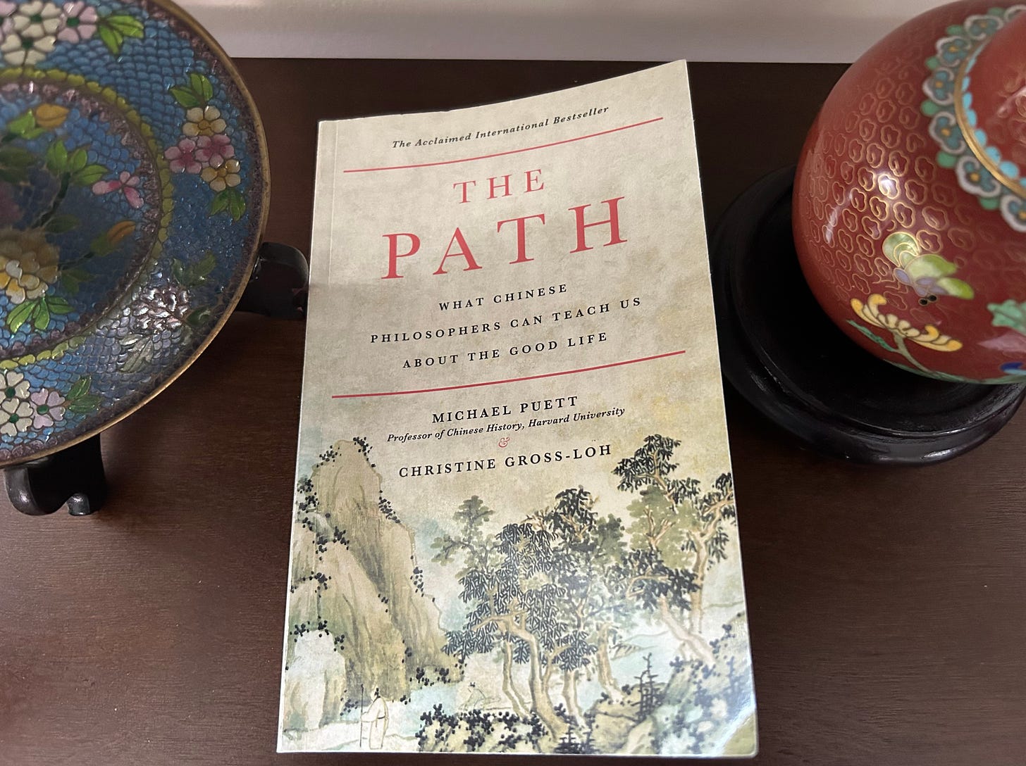 A copy of The Path sitting on a shelf in between a blue decorative plate and red urn.