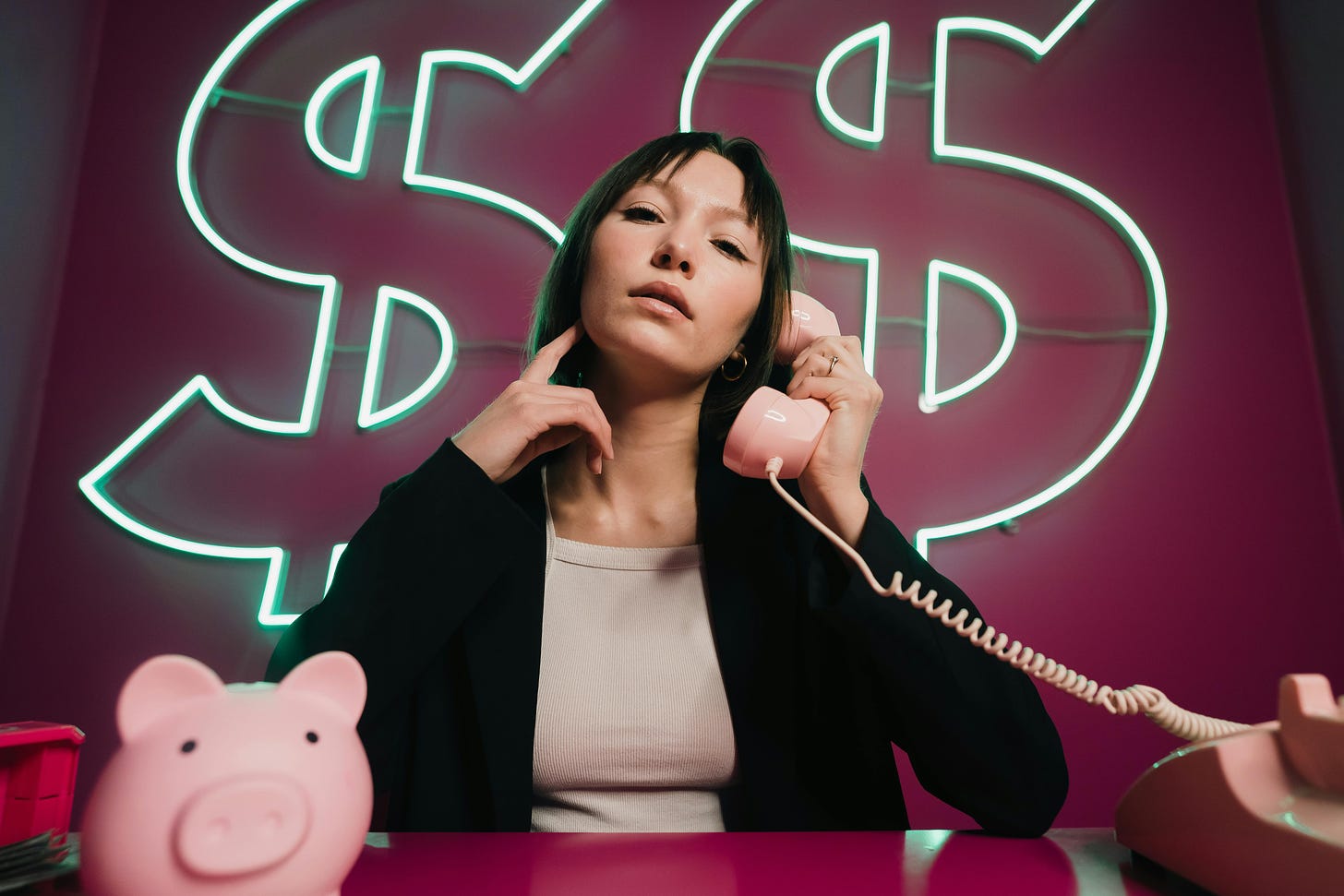 A dark-haired woman wearing a light pink shirt and black sweater sits at a dark pink desk. She is holding a pink telephone receiver in her left hand up to her ear. Behind her are two neon dollar signs on a dark pink wall. On the desk in front of her is a pink piggy bank.
