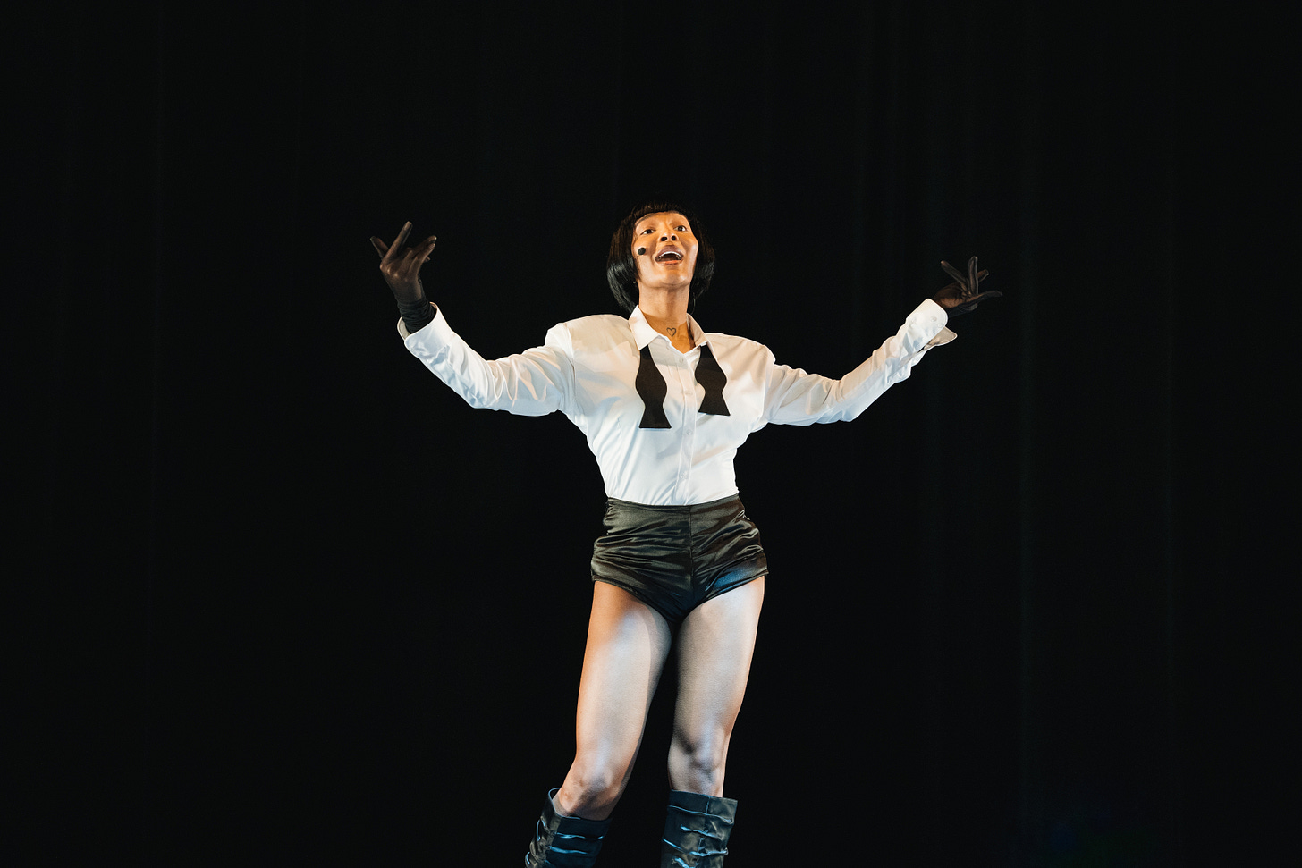 Ms. Boogie stands confidently on stage, wearing a white button-up shirt with a black bow tie, black gloves, high-waisted black shorts, and knee-high black boots. Her arms are outstretched, and she exudes an expressive energy, with her face lit up as she performs against a dark, minimal background.