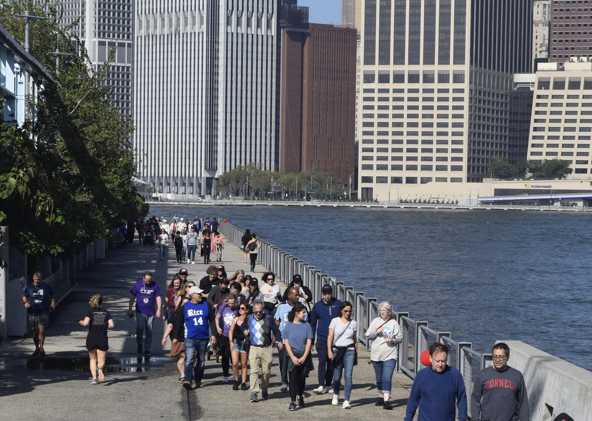 A group of people walk on a path next to a river with the Manhattan skyline in the background.