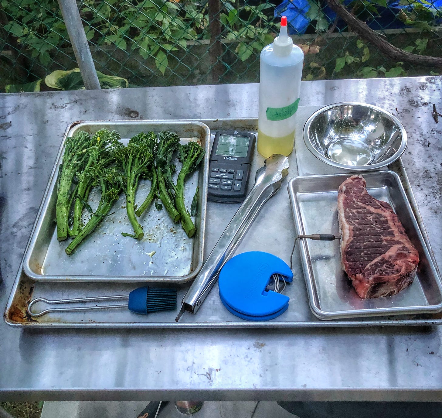 A photo of a half sheet pan set up to carry things out to grill. On the half sheet pan there is a silicone brus, a squeeze bottle of oil, a pair of tongs, an electronic thermometer. The probe of the thermometer has been inserted into a raw steak, which is sitting on an eighth sheet pan, also on the half pan. There is also an quarter sheet pan that is holding spears of broccolini that have been tossed in olive oil, salt, and pepper. 