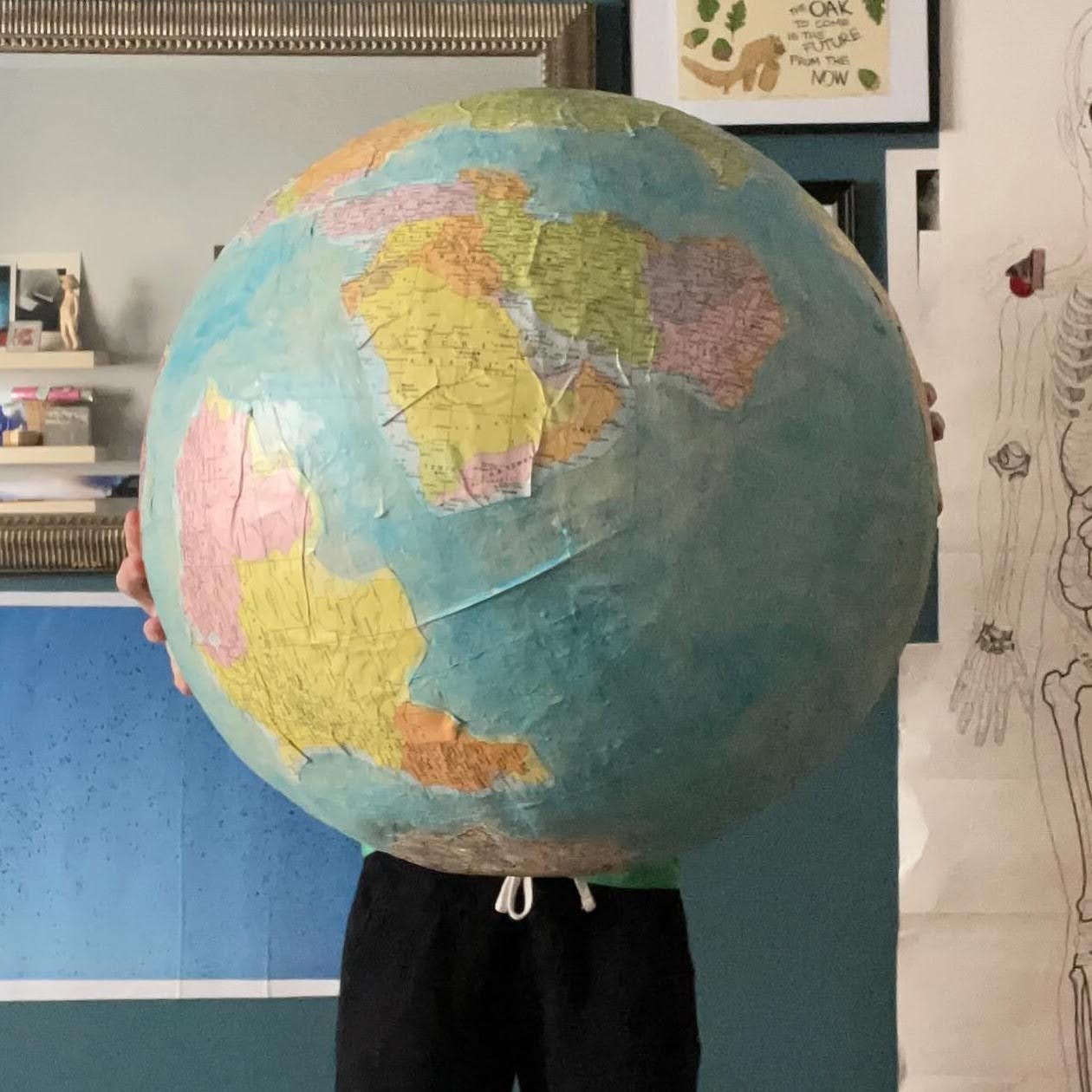 image of a white woman holding an enormous, paper mache globe pinata; the globe obscures the upper half of her body and is geographically inaccurate, with collaged imagined geographies