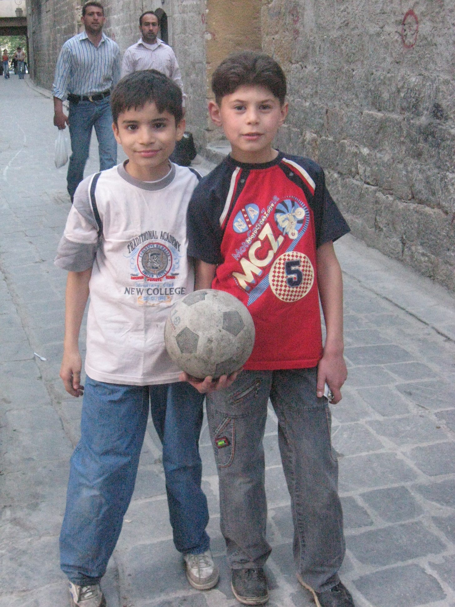 Two young Syrian boys wearing t-shirts and jeans pose on a paved brick street.. One holds a soccer ball.