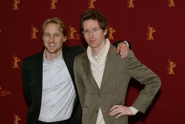 Two people pose on a red carpet. One wears a dark suit and striped shirt, the other a light-colored suit with a scarf