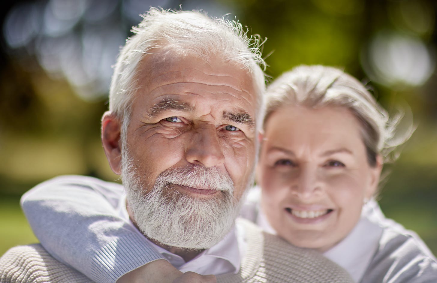 Older white man with white hair and beard looks into camera. A younger white woman in background has her right arm lovingly around his neck and is out of focus.