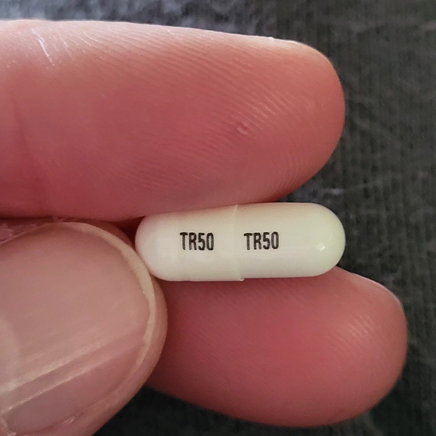 Fingers holding a tiny white medication capsule with T50 written on it in black.