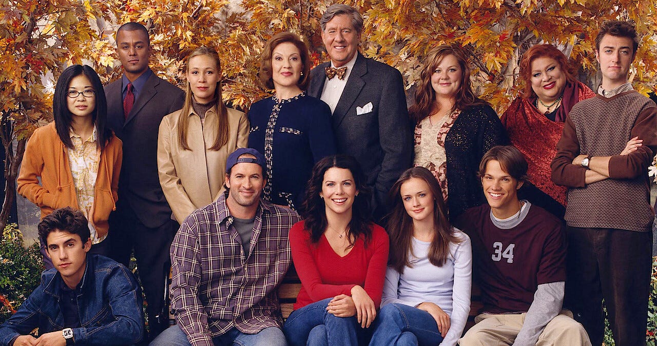 Foto com um fundo de árvores com folhas amarelo-outono e todo o elenco de Gilmore Girls na frente, talvez da segunda temporada por já envolver Jess mas ainda trazer Dean.