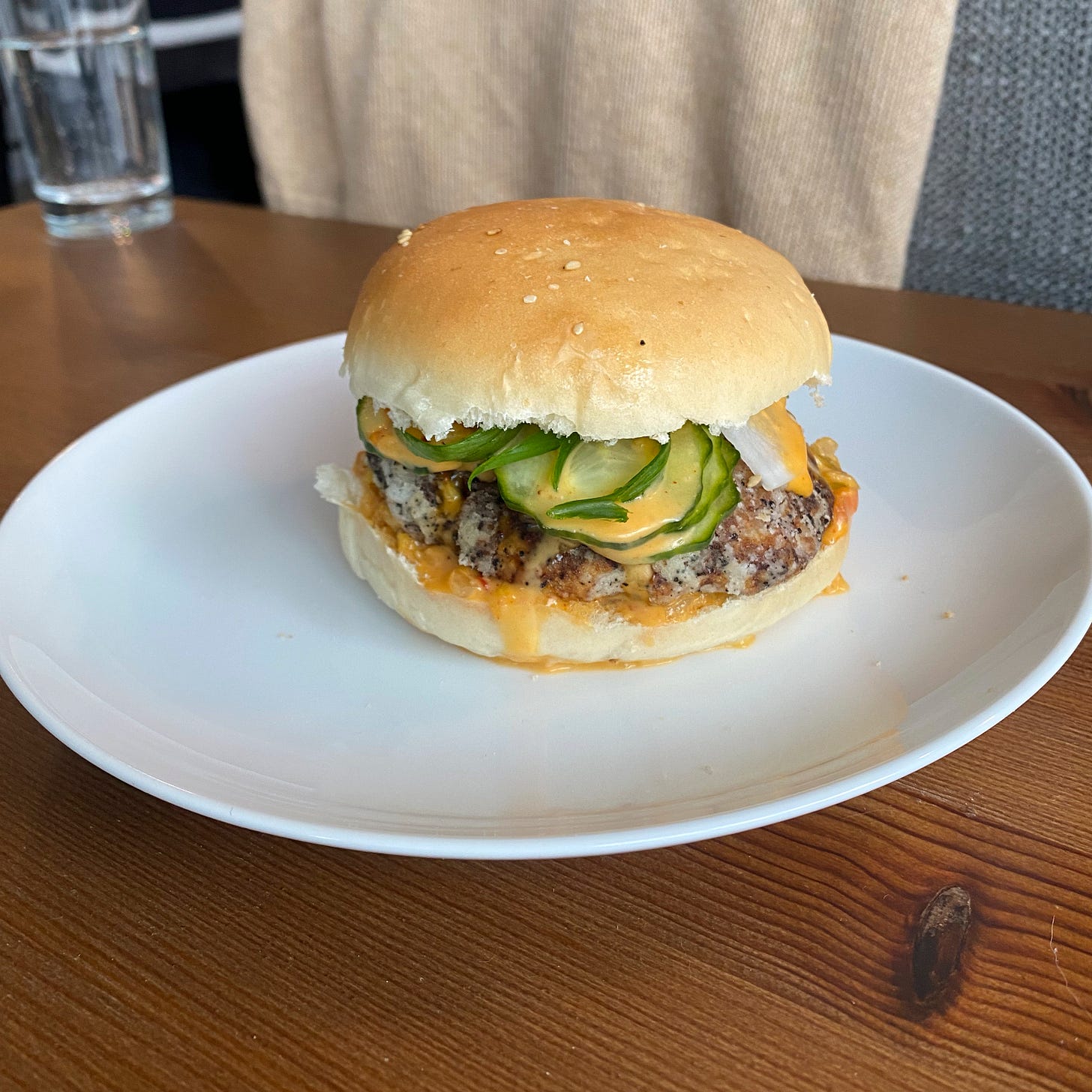 A white plate on a wooden table. On the plate is a seitan burger on a white bun, oozing with an orange sauce, and pickled cucumber and daikon are visible spilling over the edge.