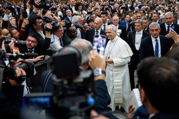 Pope Leo XIV in white vestments surrounded by news media and a large crowd of other people. Pope Leo XIV in white vestments surrounded by news media and a large crowd of other people.