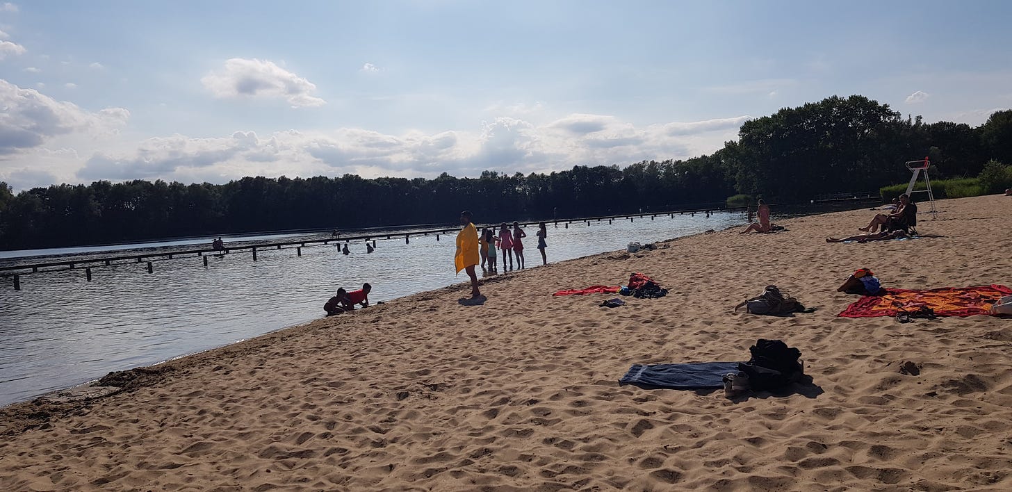 Photo of a lake on a sunny day with a sandy shore and people standing at the water's edge
