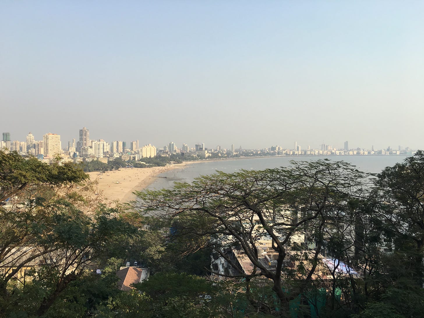 View of a smaller part of Mumbai’s skyline seen from above. Below, the beach, the tops of homes are faintly visible through trees.