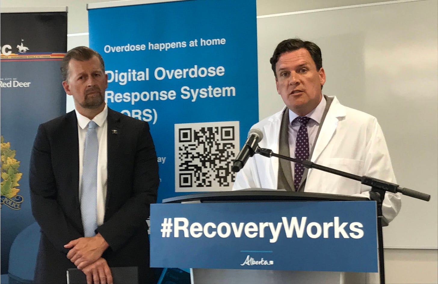 Minister of public safety Mike Ellis stands behind Dr. Nathaniel day, who speaks at the podium marked #RecoveryWorks. In Behind, A Banner reads Overdose Happens At Home, Digital Overdose Response System