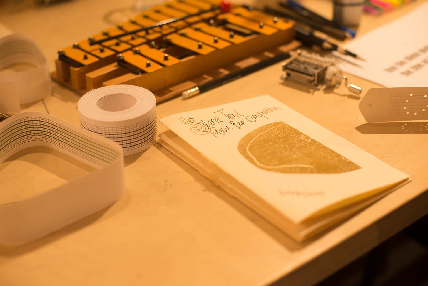 A table is filled with musical composition tools. There is a booklet titled "Slime Trail Music Box Composition," rolls of perforated music box paper, a hand-crank music box, a xylophone, and various pens and pencils, suggesting a workspace for creating music box compositions.