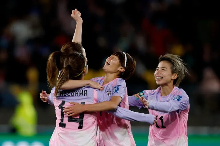 On a soccer field, four Japanese women players in pink-and-lavender uniforms, celebrate in a group hug, with one of them raising a fist into the air.