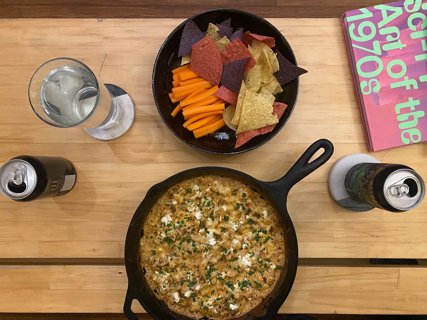 A coffee table from above, showing a black shallow dish with tri-colour tortilla chips and bright orange carrot sticks. In front of it is a cast iron pan of the buffalo dip described above: orange-brown with flecks of white feta and a dusting of chopped parsley. Surround the dishes are two cans of beer, a glass of water, and a coffee table book with a pink/purple cover and lime green text entitled 'Sci-Fi Art of the 1970s'.