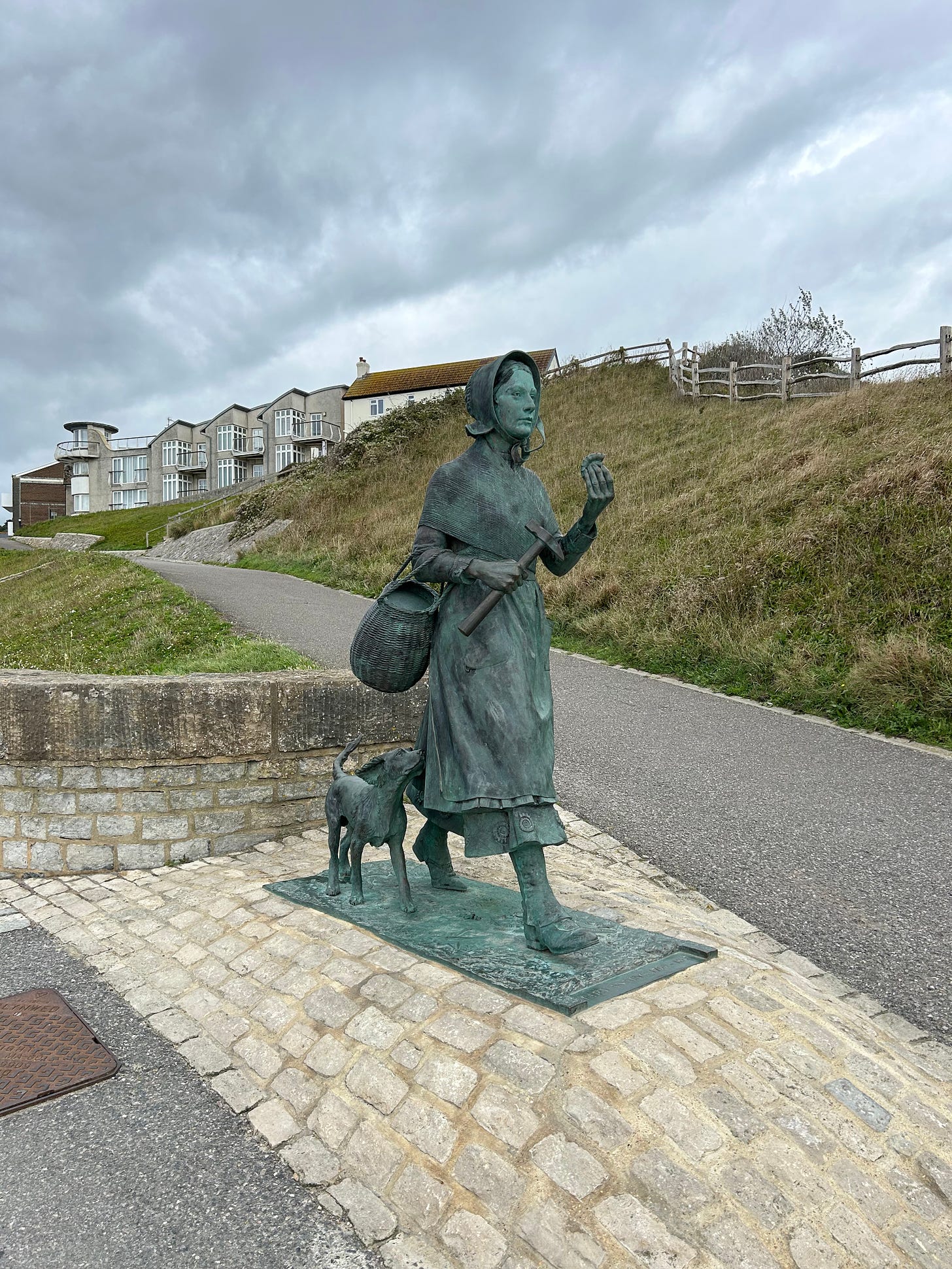 A bronze statue of Mary Anning and her dog. She is looking out to sea across a bay not in view in this photo. In the background is a grassy bank and some houses on the horizon. Image: Roland’s Travels