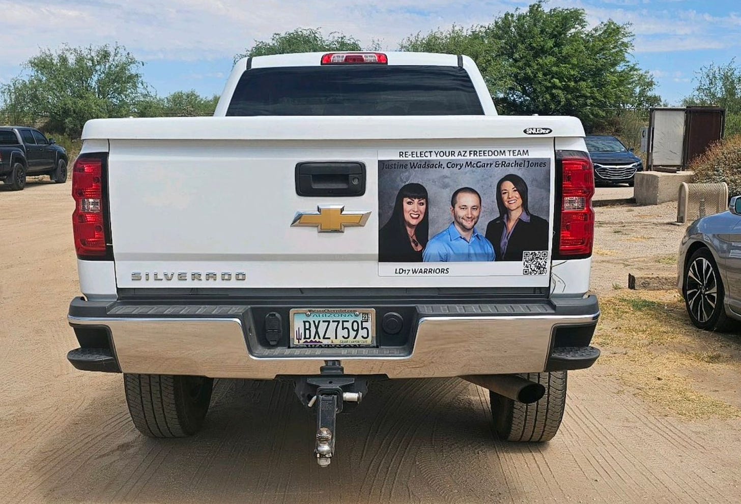 A white Chevy Silverado truck parked in a dirt parking lot. A large sticker taking up half their tailgate is an image of Justine Wadsack, Cory McGarr, and Rachel Jones, aka the “AZ Freedom Team.” The bottom text reads “LD17 warriors.” AZ plates read BXZ7595.