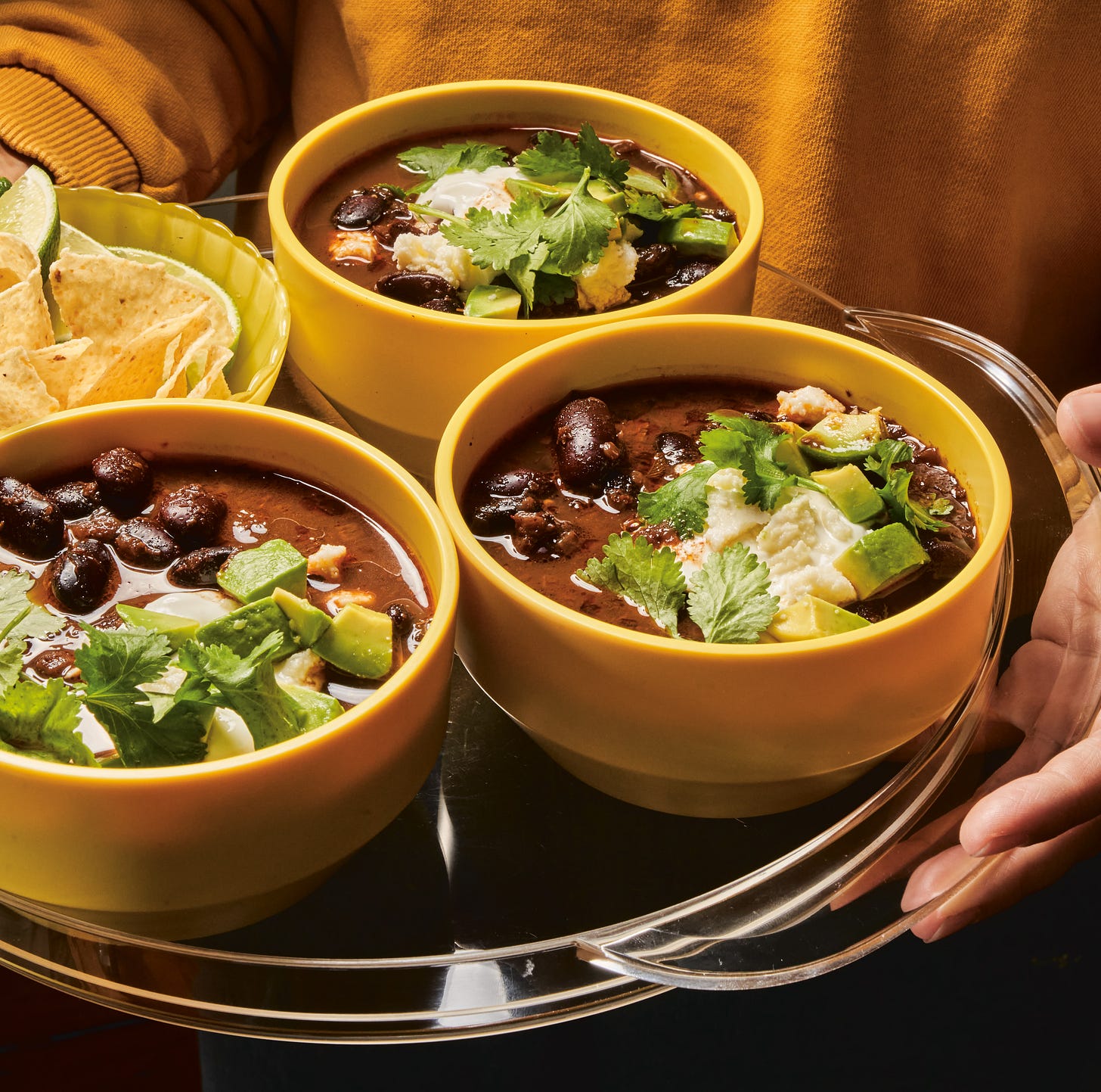 3 yellow bowls of black bean soup with avocados and cilantro, on a tray.