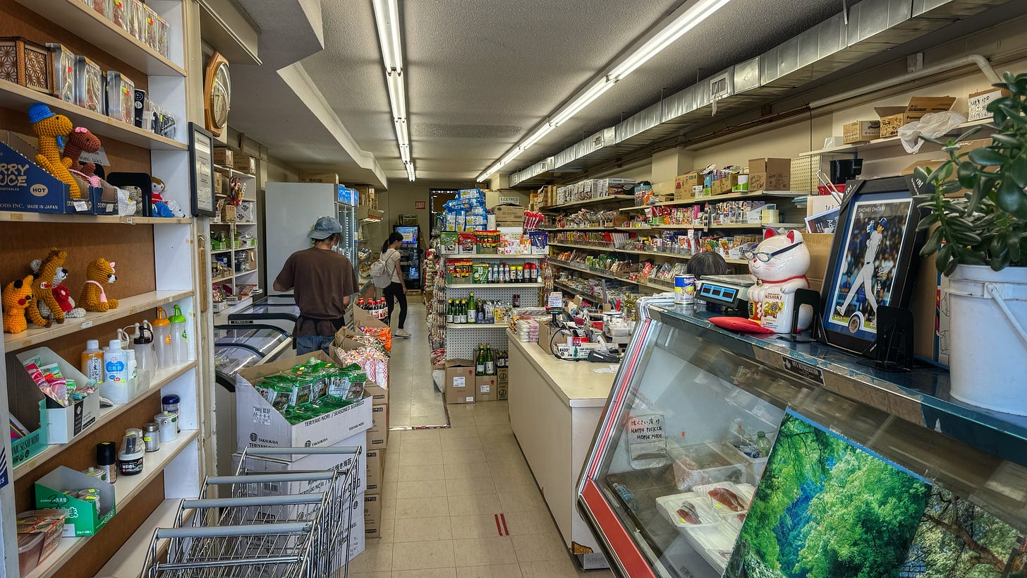 A photo taken from just inside the front door of Sandown Market. On the left side there is a series of shelves, fridges, and freezers. On the right there are a refrigerated display case, a cash register, and, farther back, more shelves. Everything is very plain and slightly cluttered.