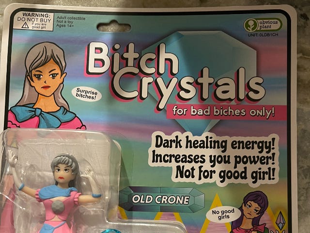 Photo of a toy package with the words Bitch Crystals. For bad bitches only. Dark healing energy! Increases your power! Not for good girl. Includes an action figure of a white woman with long gray hair wearing a pink dress with a blue ribbon.