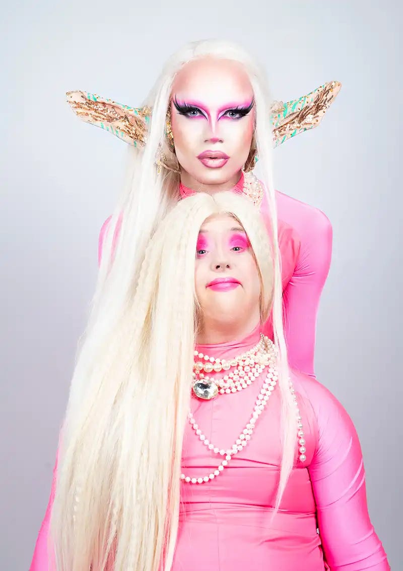 Two artists, one with Down Syndrome, look like sisters, made up in high femme drag. They have platinum blonde hair, tight and bright pink bodysuits, and shockingly pink makeup.