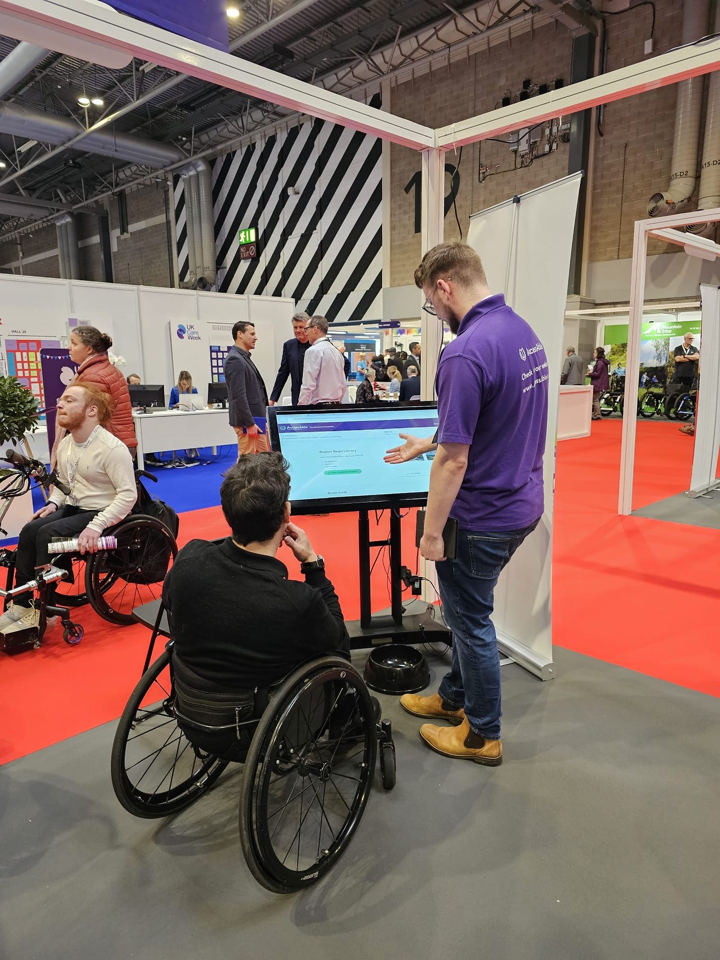 A male member of AccessABle demonstrates how to use the detailed access guides on a touch screen to a visitor to the Naidex stand 