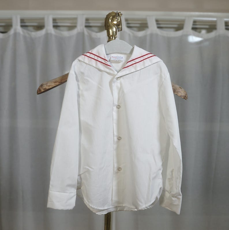 May include: A white long-sleeved shirt with a red trim sailor collar. The shirt has a button-up front and long sleeves with button cuffs.