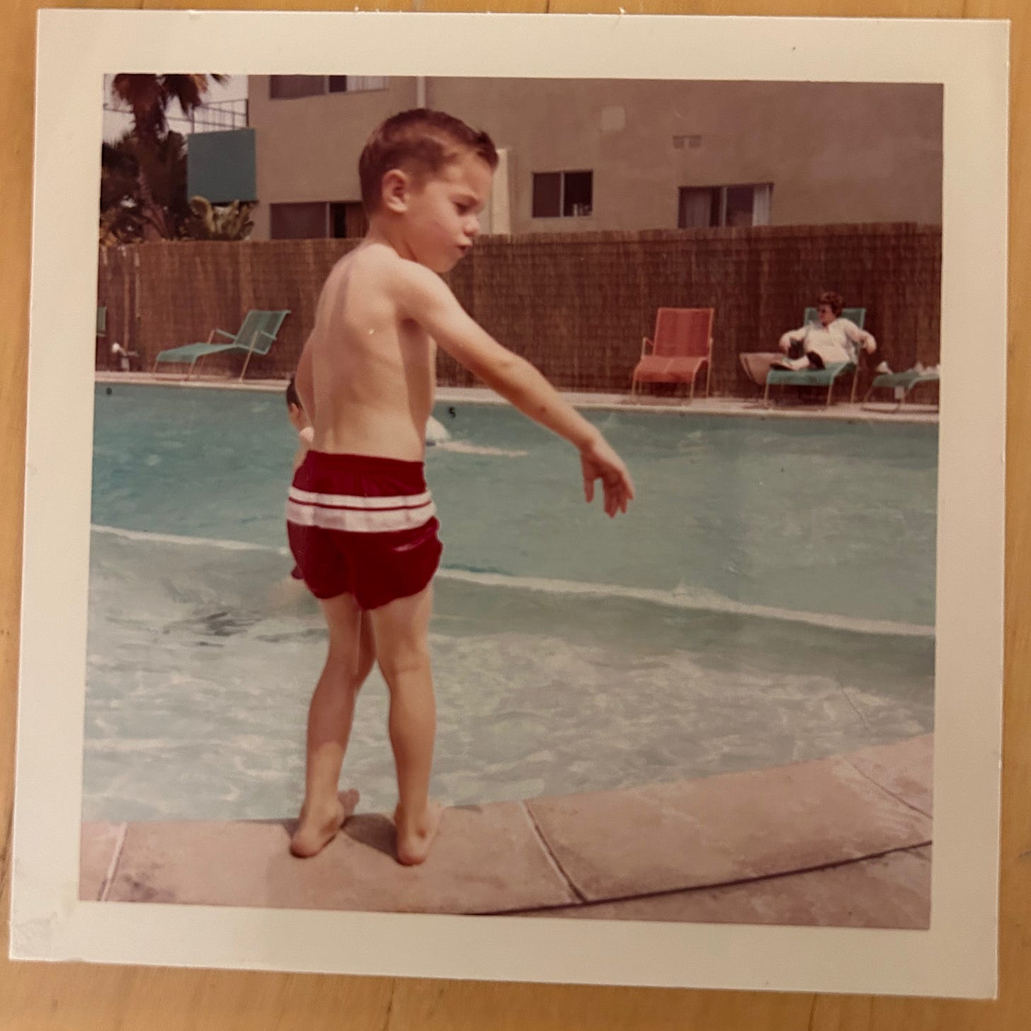 A boy maybe 6 years old is standing at the edge of an outdoor pool with his hand outstretched. The photo seems to have been taken in the 60's.