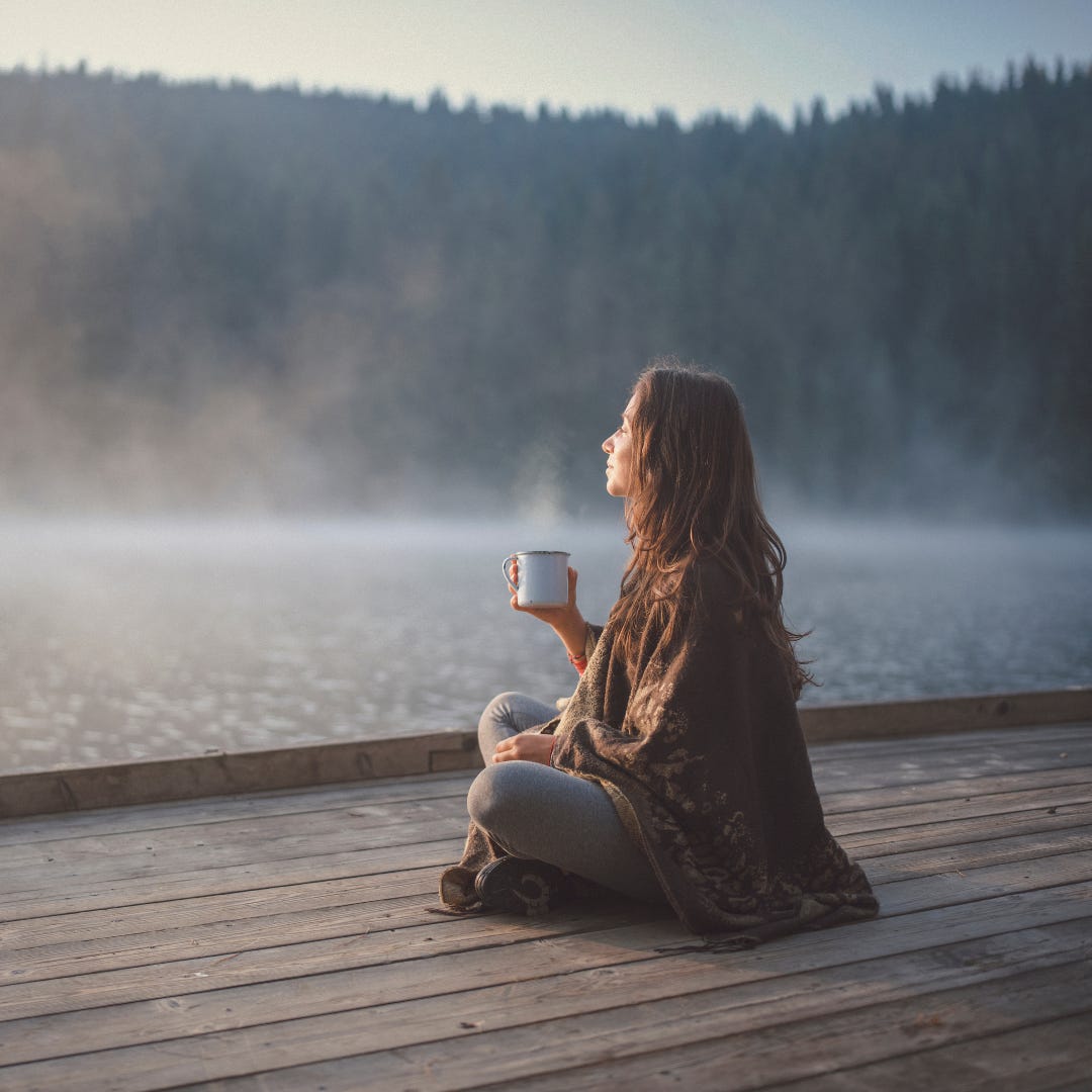 A woman with a steaming mug in one hand, sitting cross legged and eyes closed on a boardwalk by a lake with tree covered mountains in the foggy distance.