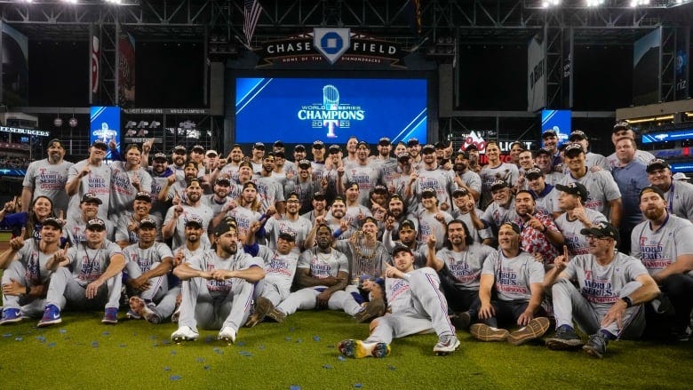 A male baseball smile while sitting together on the field for a team picture with a trophy in the middle.