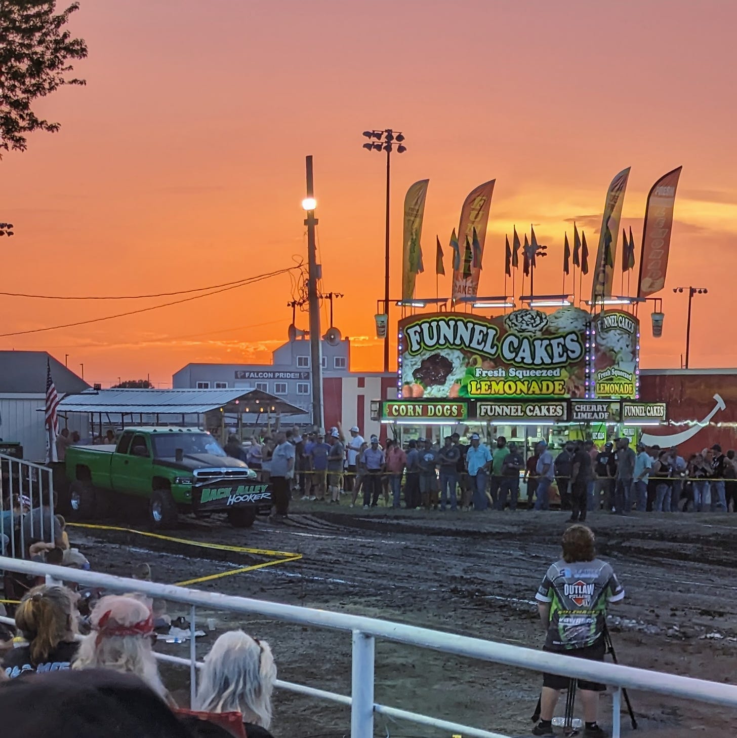 A green truck getting ready to pull a weighted sled across a dirt field. A funnel cake stand is behind it, and the sun it setting.