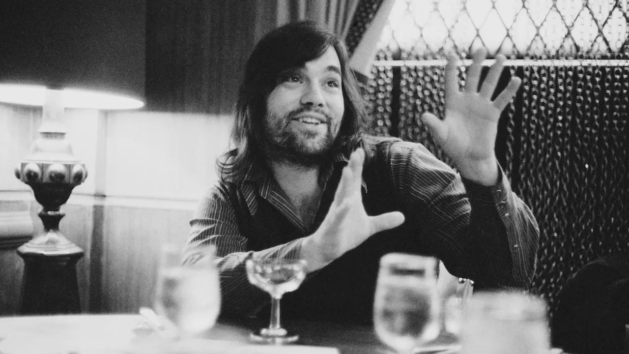 Lowell George sitting at a table holding his hands up in an enthusiastic gesture.