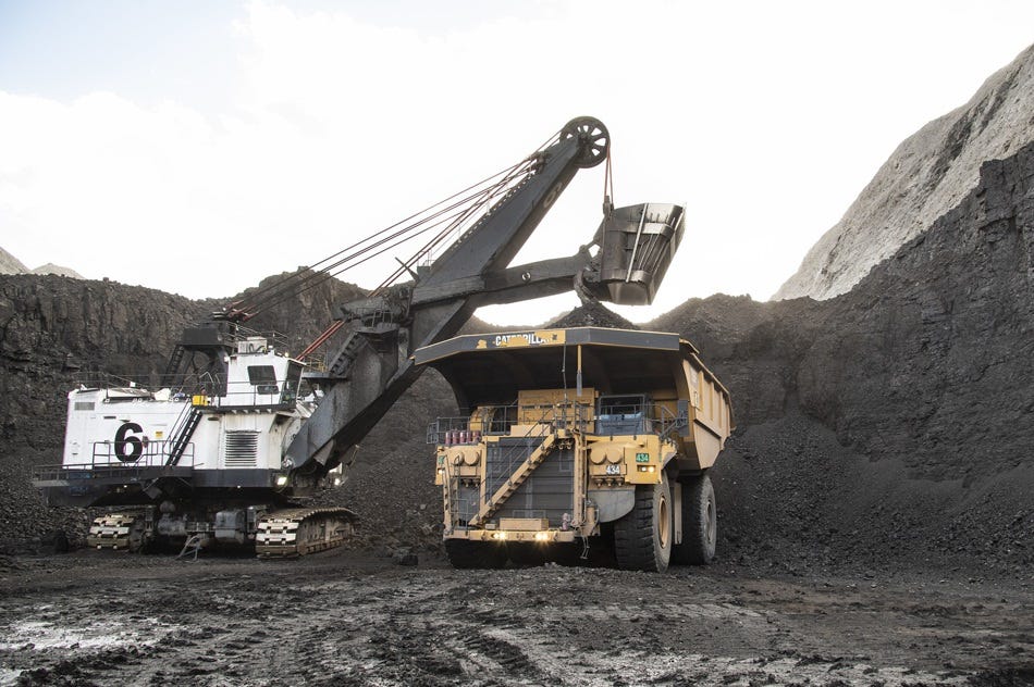 A giant white power shovel loads a giant yellow dump truck deep in an open pit mine, under a cloudy sky