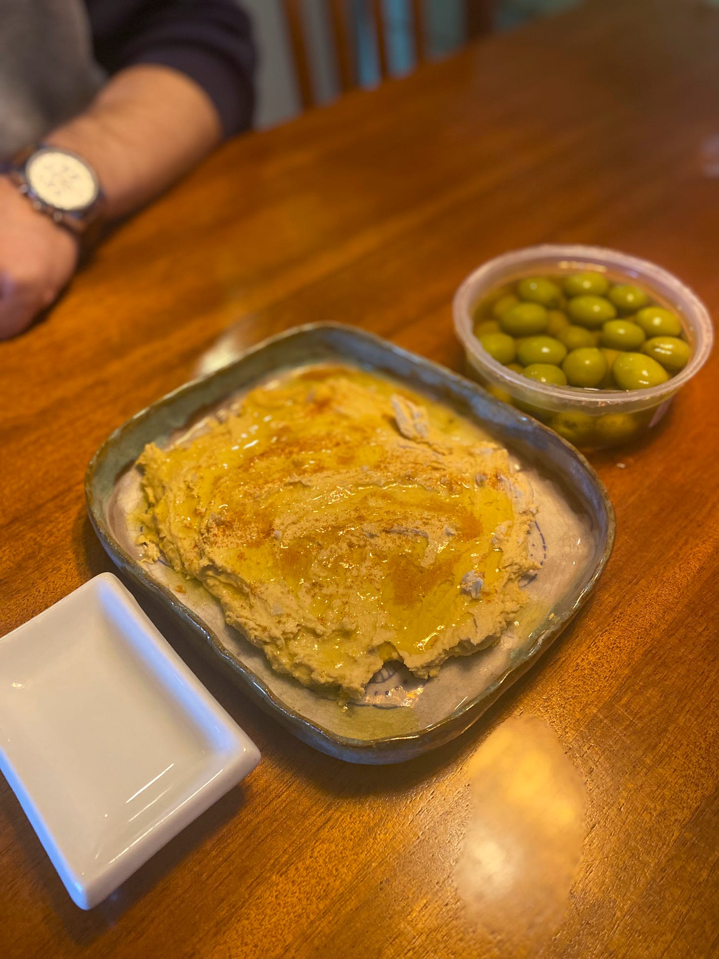 A squarish handmade dish spread with hummus that is covered with olive oil and paprika. On one side of the dish is a container of castelvetrano olives, and on the other side is a small white dish for the pits.