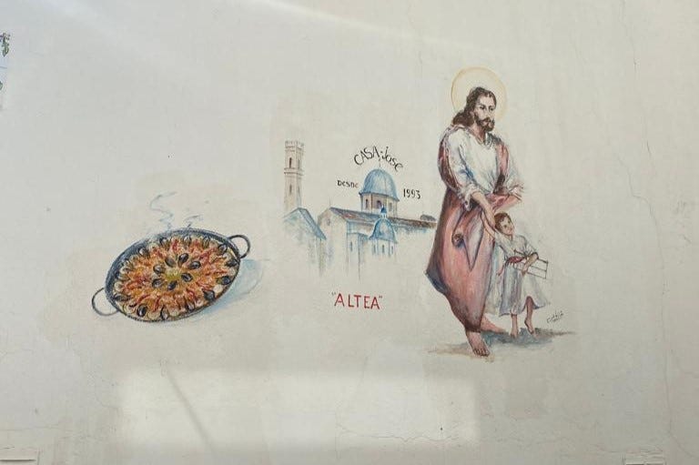 a painting on a wall of a pan of paella, the city of Altea, and Jesus with a little boy or angel