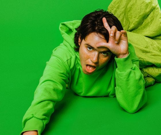 An image of An(Dre)a a person with dark hair wearing a green hoody lying down putting an L above their head. The background is also green. 