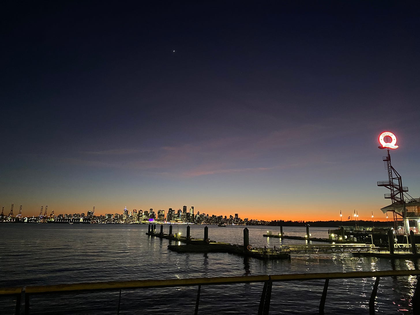 A photo of Vancouver at dusk, taken from the Shipyards in North Van. The sky is orange at the horizon, fading to yellow, light blue, and then dark blue at the top of frame. Venus is prominent right over the city skyline. In the foreground the water is lightly rippled along the pier, and the Q of Lonsdale Quay is lit up on the right side of the frame.