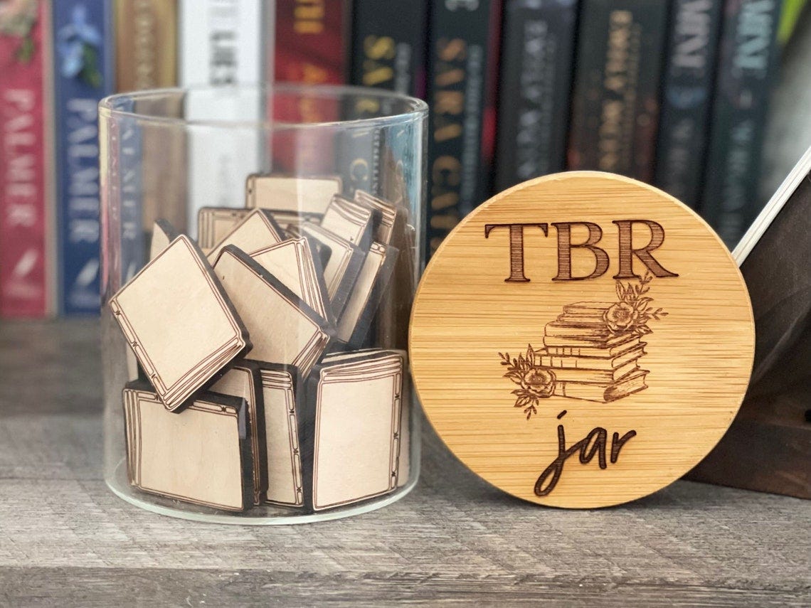 May include: A clear glass jar filled with small wooden book-shaped pieces. The jar has a wooden lid with the text 'TBR jar' and a drawing of a stack of books with flowers. The jar is sitting on a wooden surface.